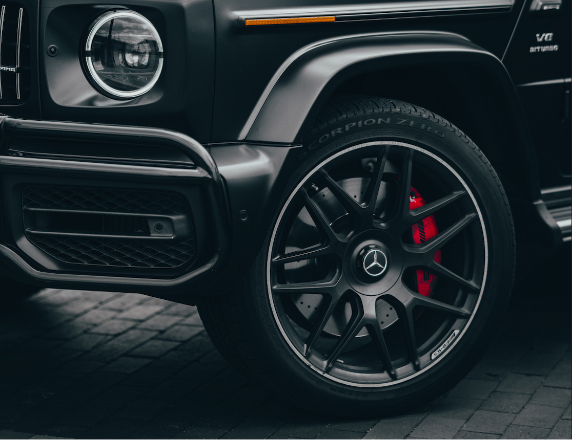 Close-up of a matte black Mercedes-Benz G-Class vehicle showing the front wheel, black multi-spoke AMG wheel, red brake caliper, and dark paved ground.